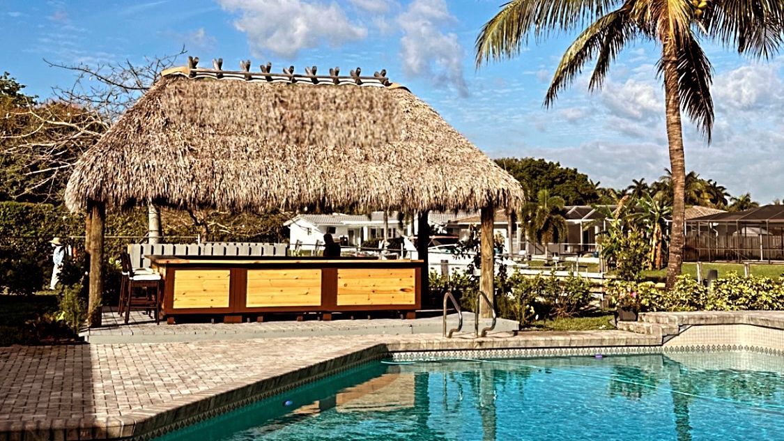 Thatched poolside tiki bar with yellow counter beside a blue swimming pool under palm trees
