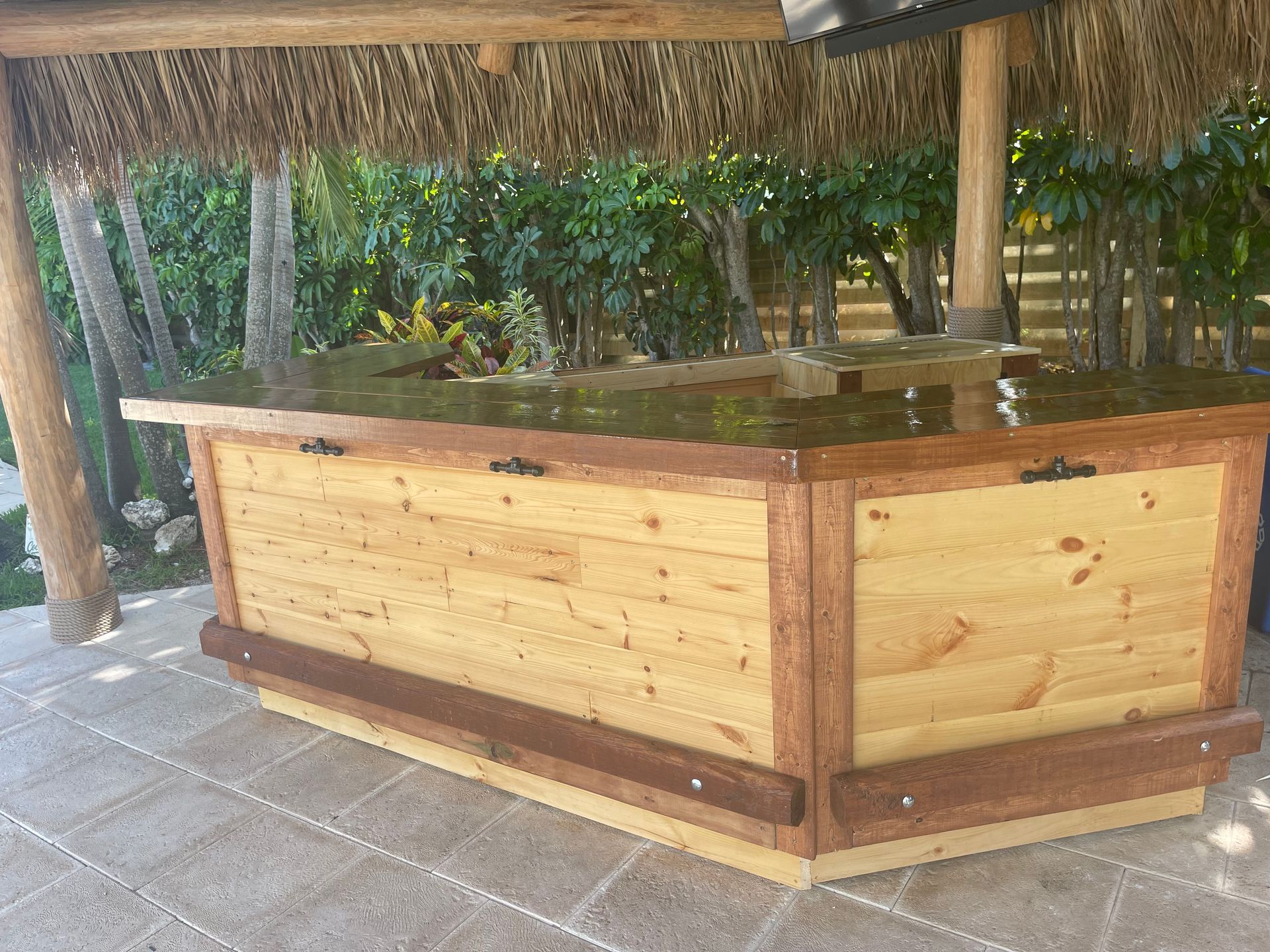 Wooden hot tub or planter under a thatched gazebo on a tiled patio, with greenery behind