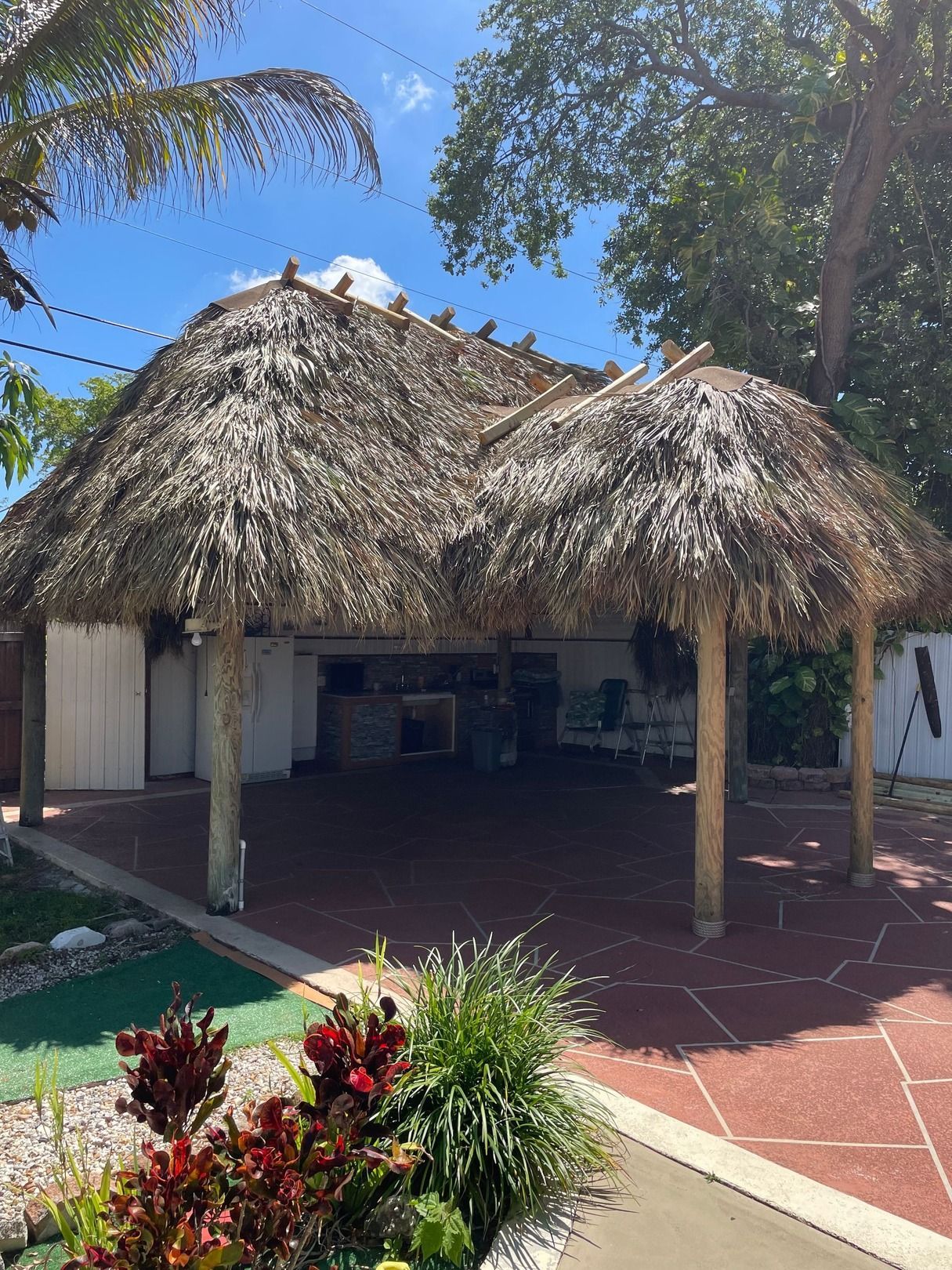 Thatched outdoor hut with tables on a sunny patio, surrounded by tropical plants and trees