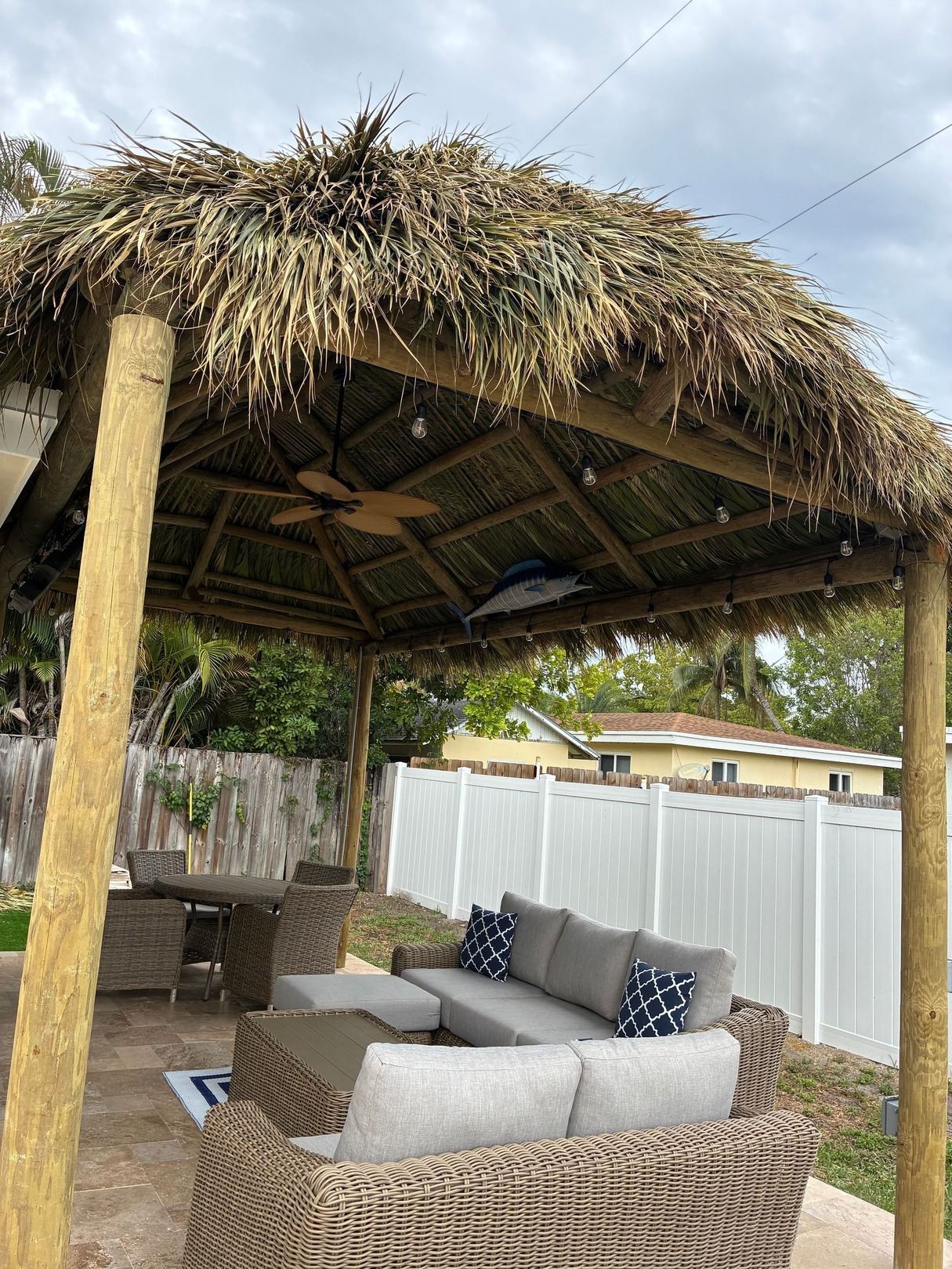 Thatched outdoor patio with wicker seating, a table, and white fence in a backyard