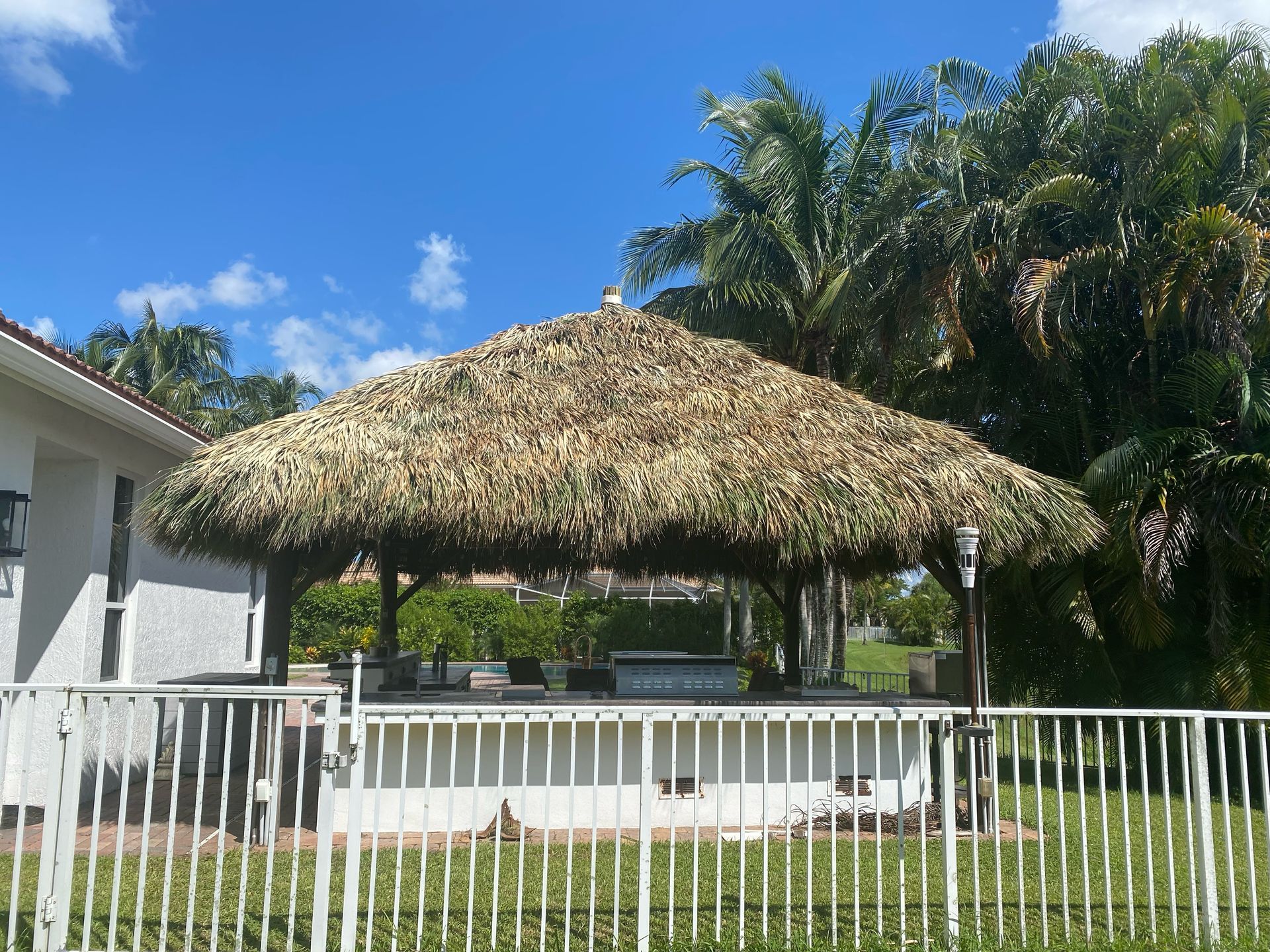Backyard tiki hut pavilion by a white fence, with palm trees and blue sky