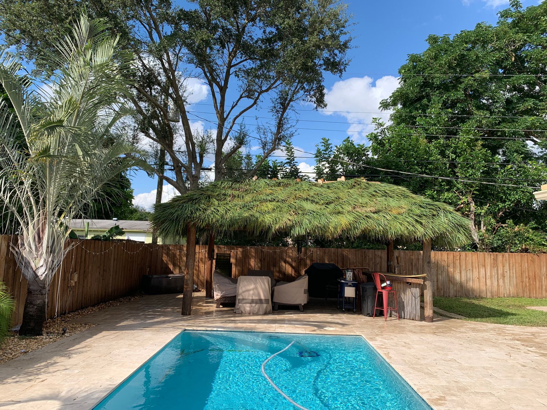 Backyard pool with thatched cabana, wooden fence, and tall trees under a blue sky