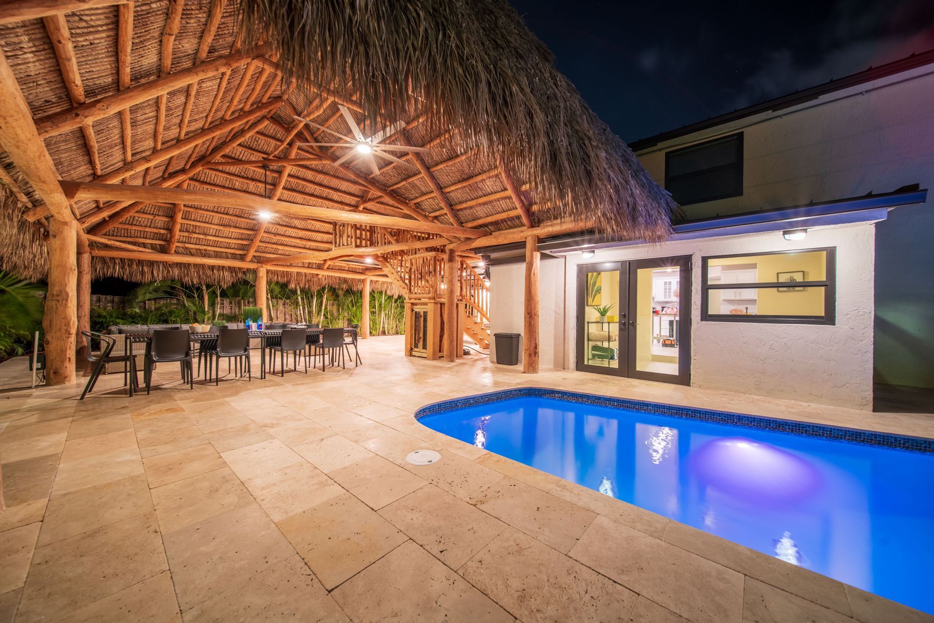 Tropical patio with a lit swimming pool, thatched roof dining area, and house at night