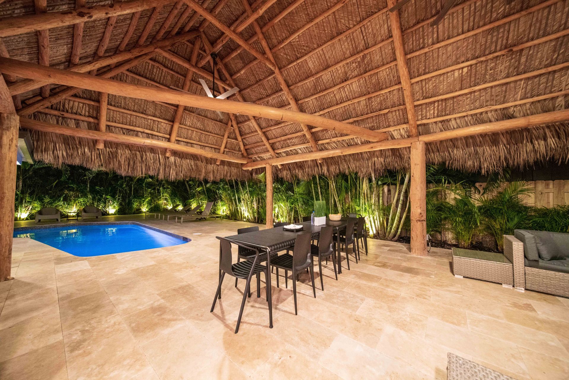 Thatched-roof patio with dining table, bar stools, lounge sofa, and a blue pool in the background