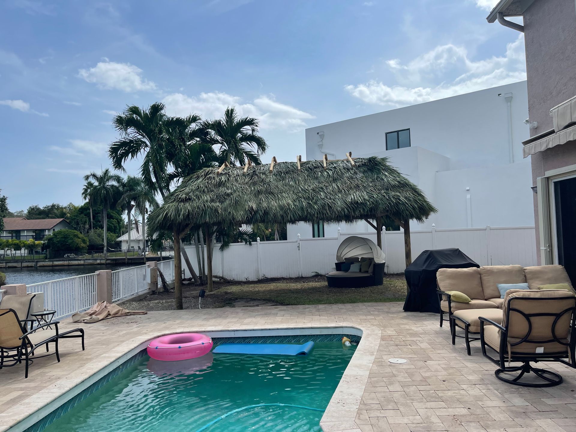 Backyard pool with pink float, lounge chairs, cabana, and white fence under a partly cloudy sky