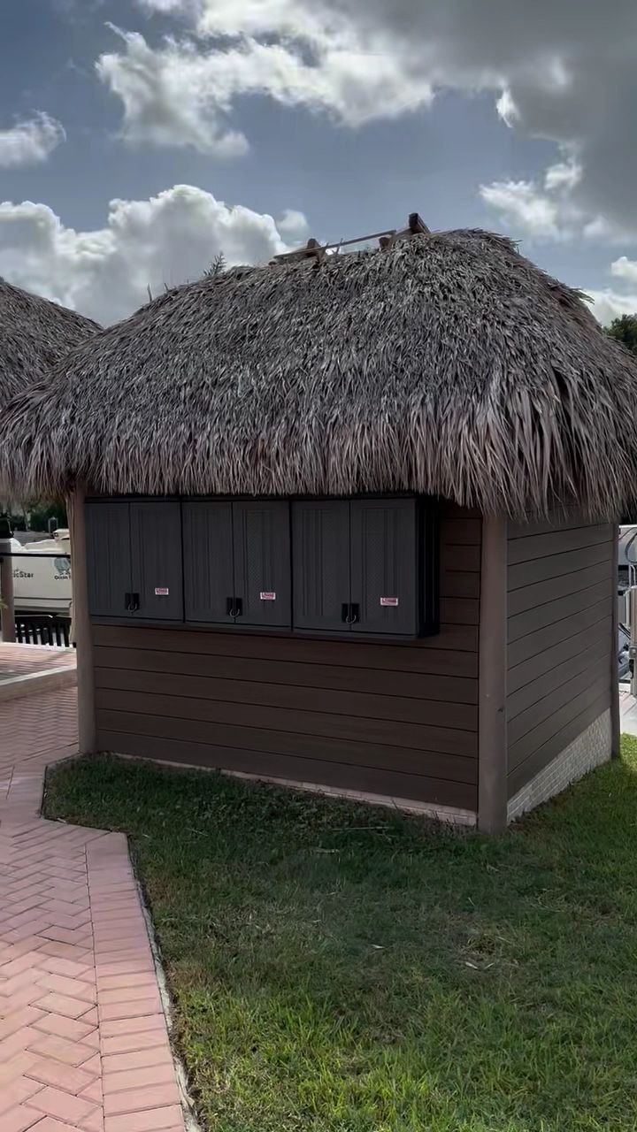 Small thatched-roof kiosk with dark shuttered windows beside a brick path and grass under a cloudy sky