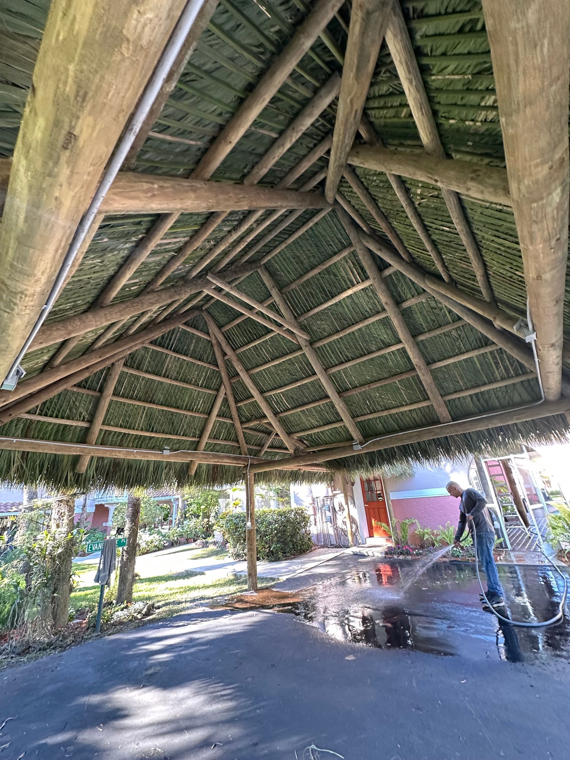 Thatched-roof pavilion with wooden beams and open sides, casting shade over a paved area outdoors