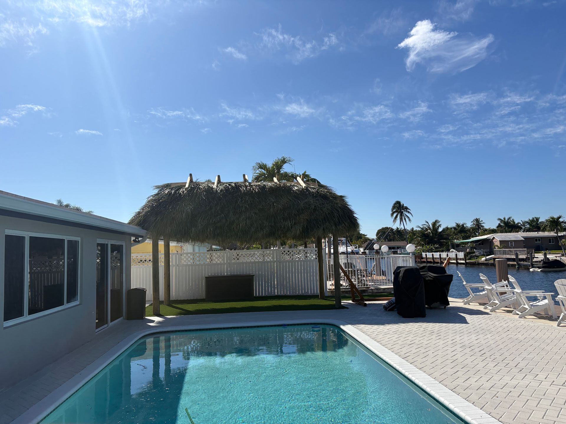 Poolside patio with cabana, white lounge chairs, and blue sky beside a house