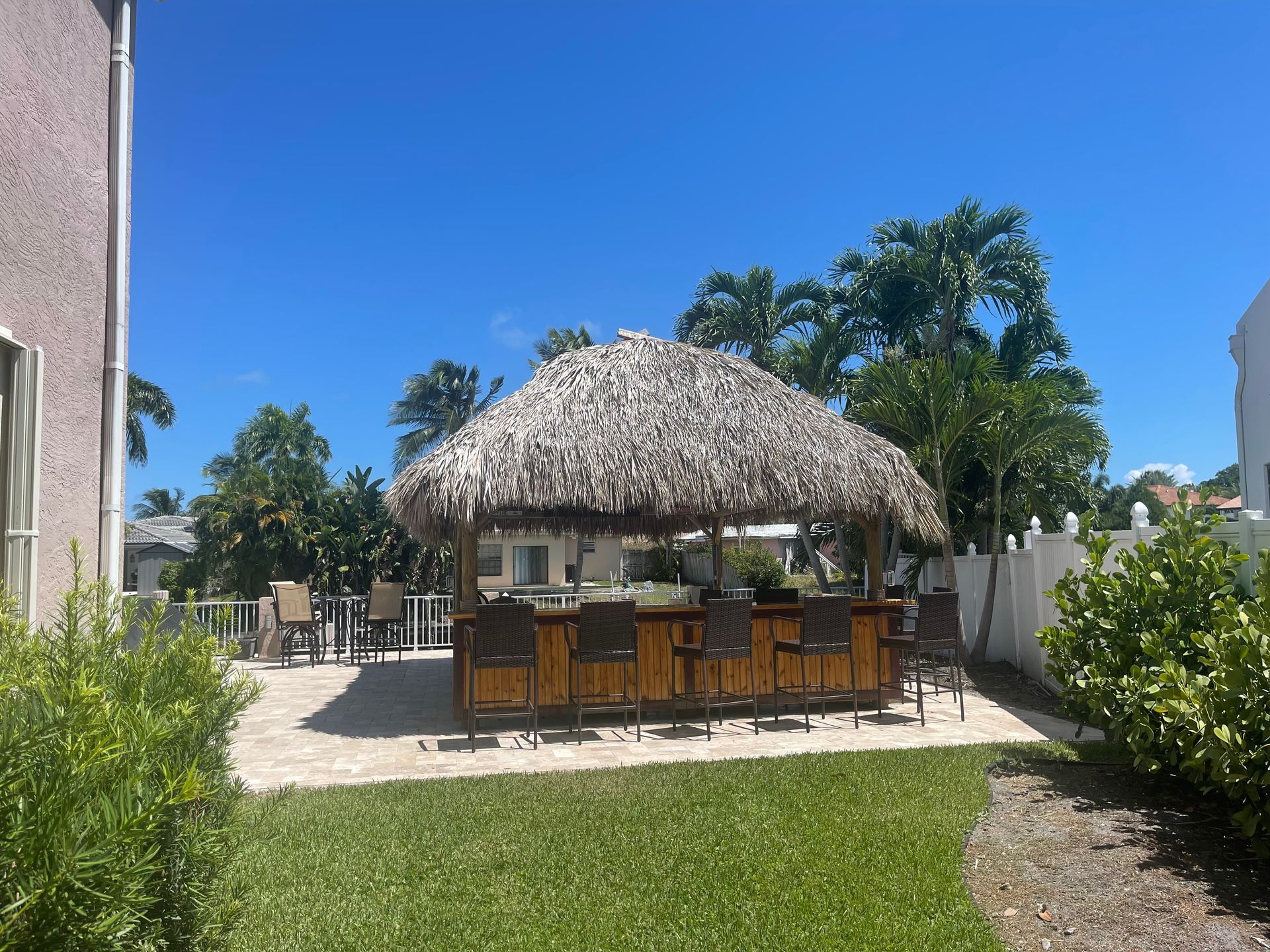 Backyard patio with a thatched-roof bar, stools, lounge chairs, and lush greenery under a bright blue sky