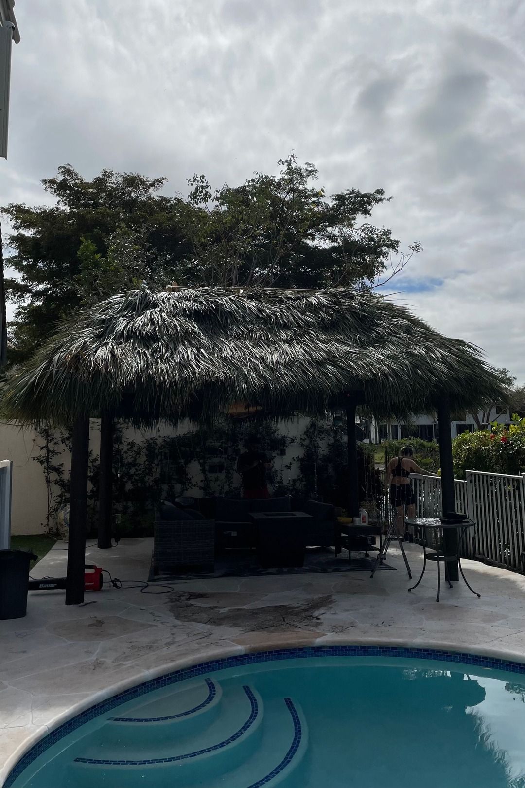 Backyard pool beside a thatched patio cabana under a cloudy sky