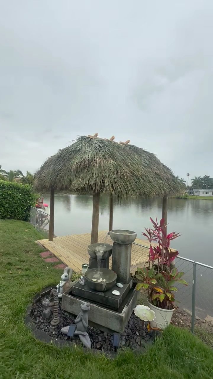 Thatched gazebo by a lakeside with a small stone altar and potted plants on the grass