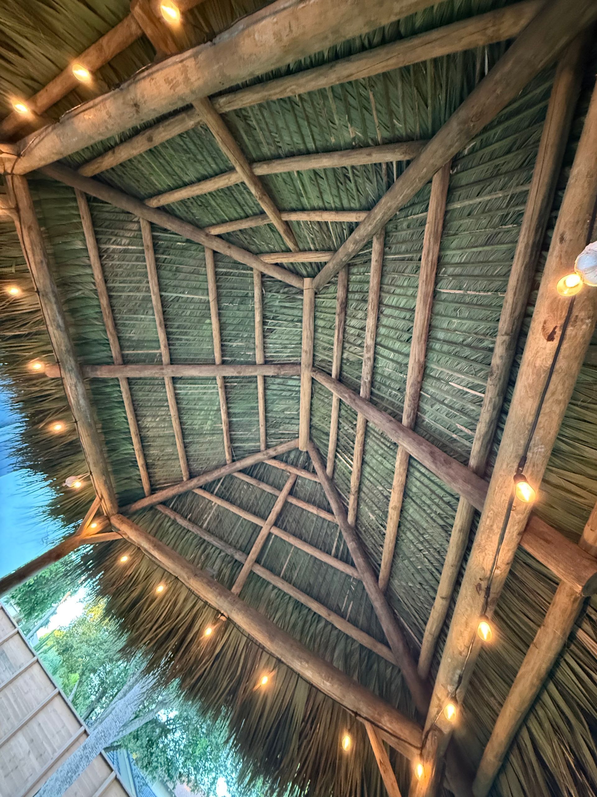 View up to a thatched pavilion ceiling with wooden beams and warm string lights at dusk