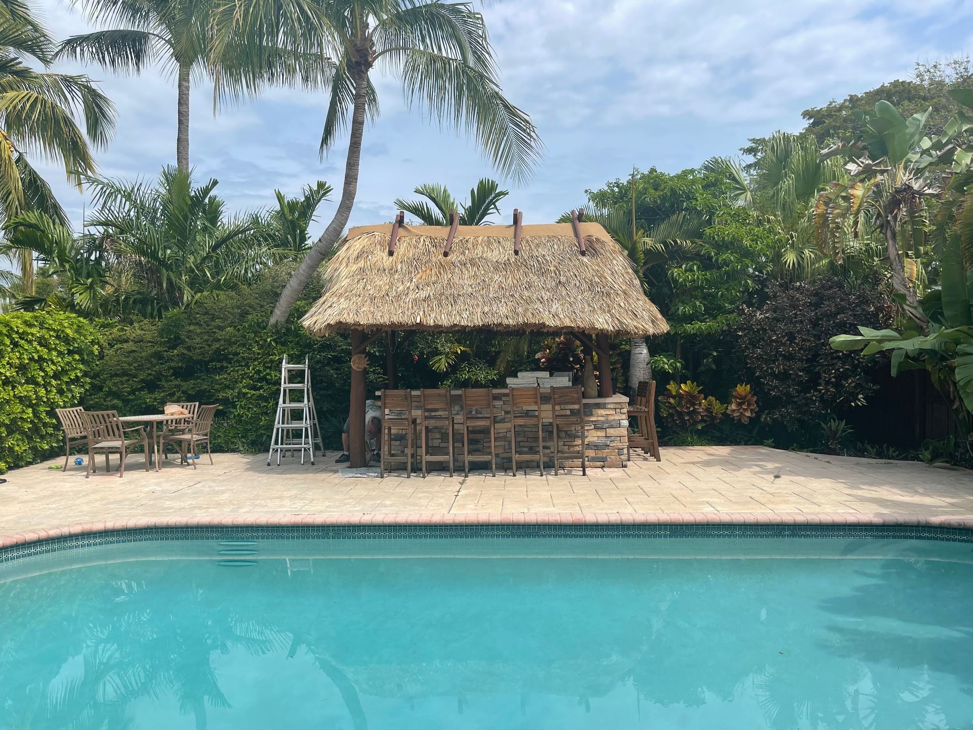 Tropical poolside patio with thatched cabana, table and chairs, and palm trees behind a turquoise pool