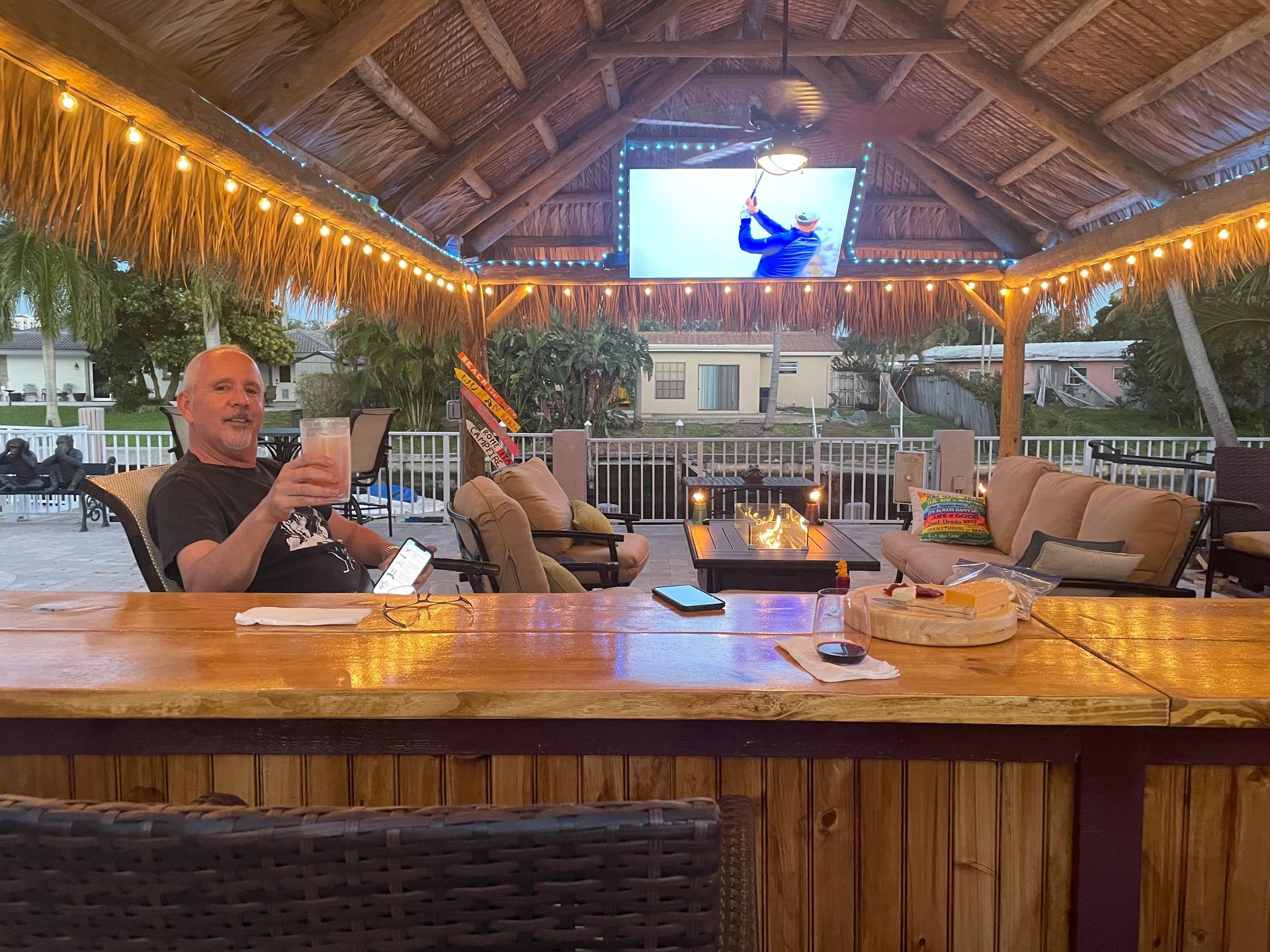 Tropical bar with a man seated at the counter under a thatched roof, lit by warm string lights and a TV overhead