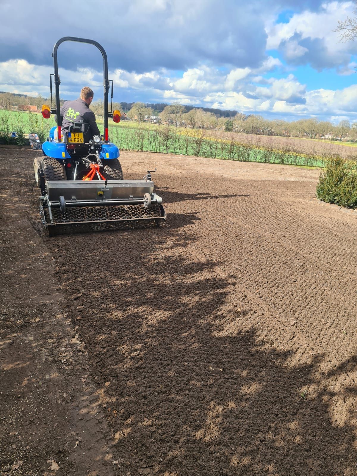 Een persoon bestuurt een blauwe tractormaaier op een veld onder een bewolkte hemel.