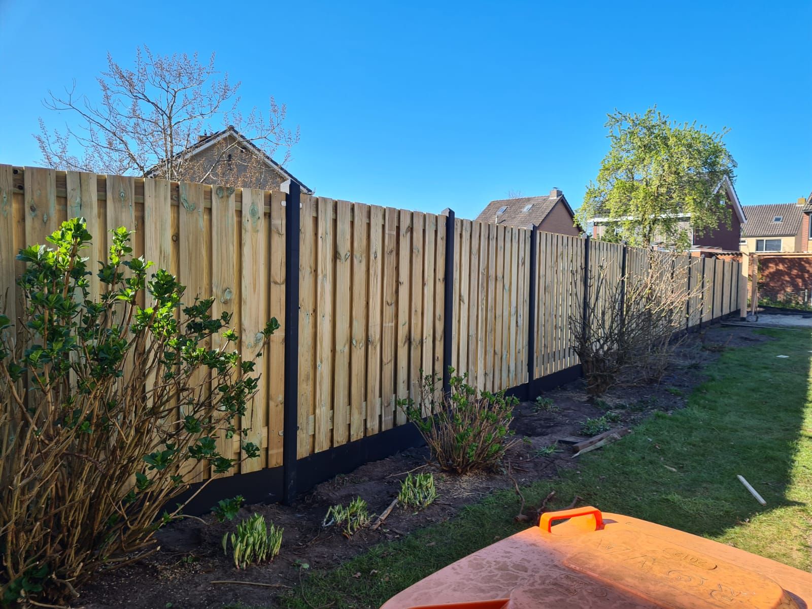 Een houten schutting langs een smal tuinpad naast een grasveld onder een helderblauwe hemel.