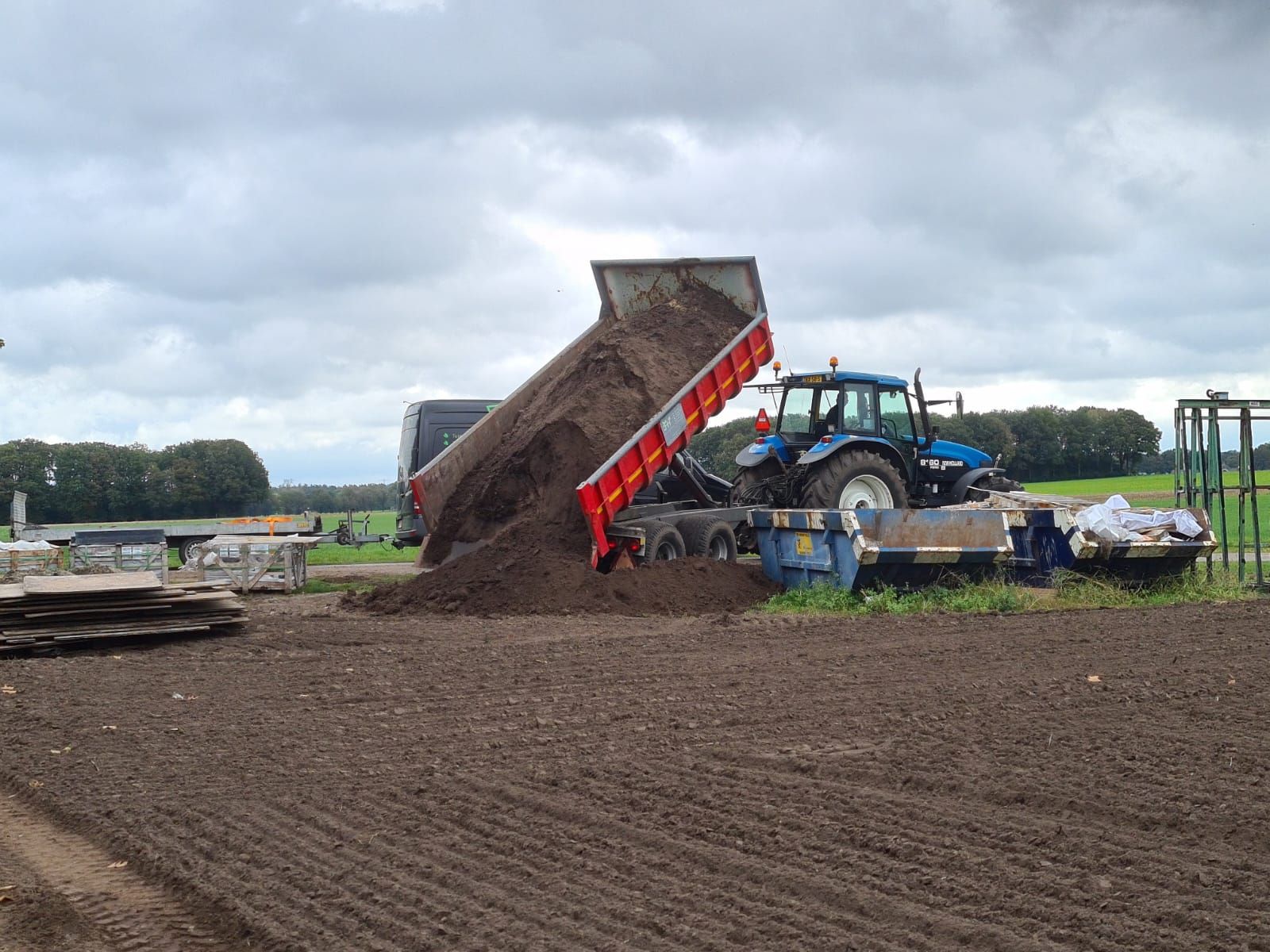 Een blauwe tractor stort grond van een verhoogde aanhanger op een modderig veld onder een bewolkte hemel.