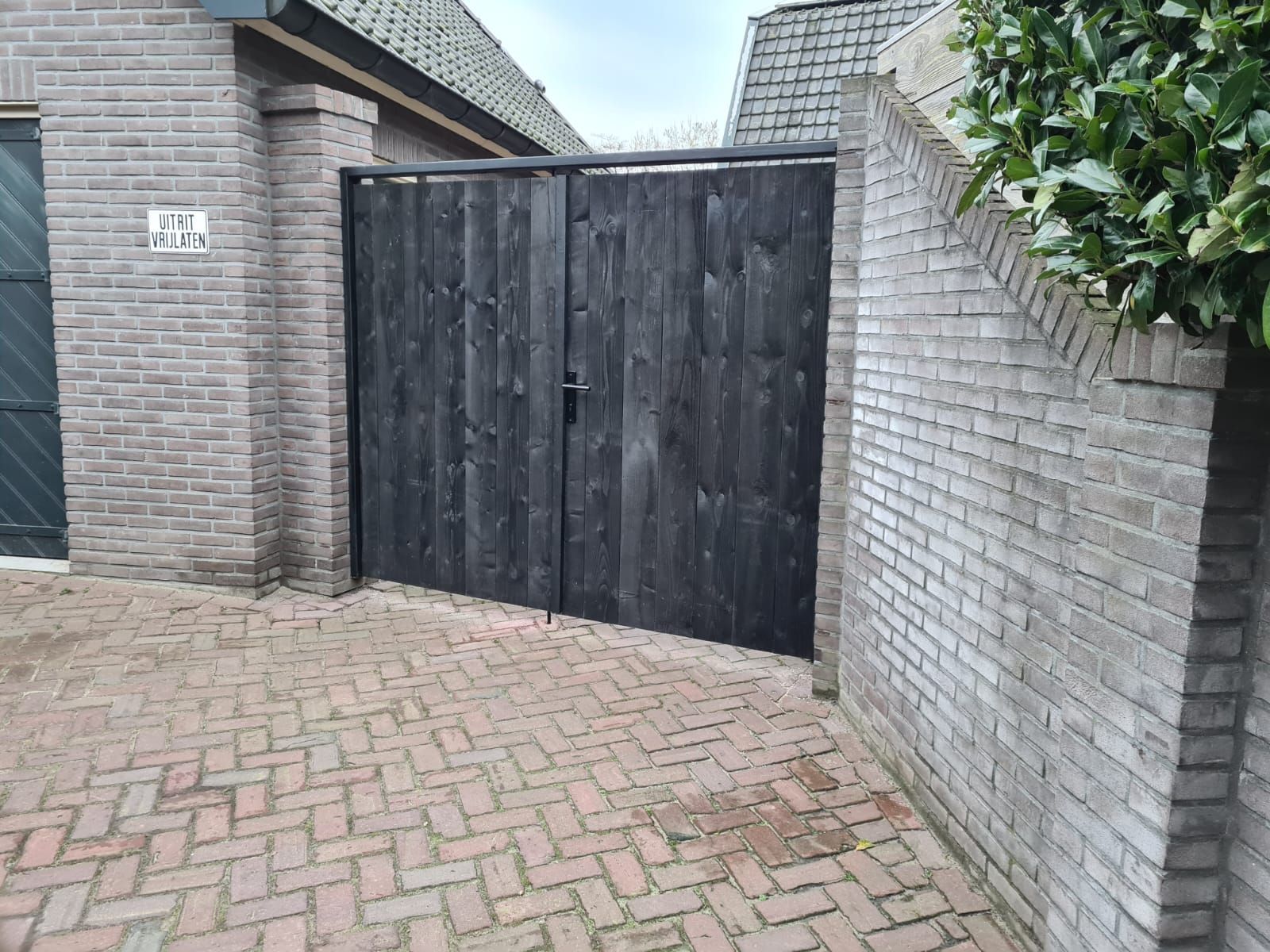 Zwarte houten poort tussen bakstenen muren naast een geplaveide oprit.