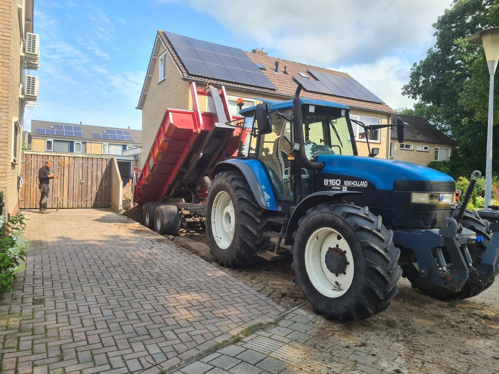 Een blauwe tractor kantelt een rode aanhanger op een bakstenen oprit naast een huis en garage.