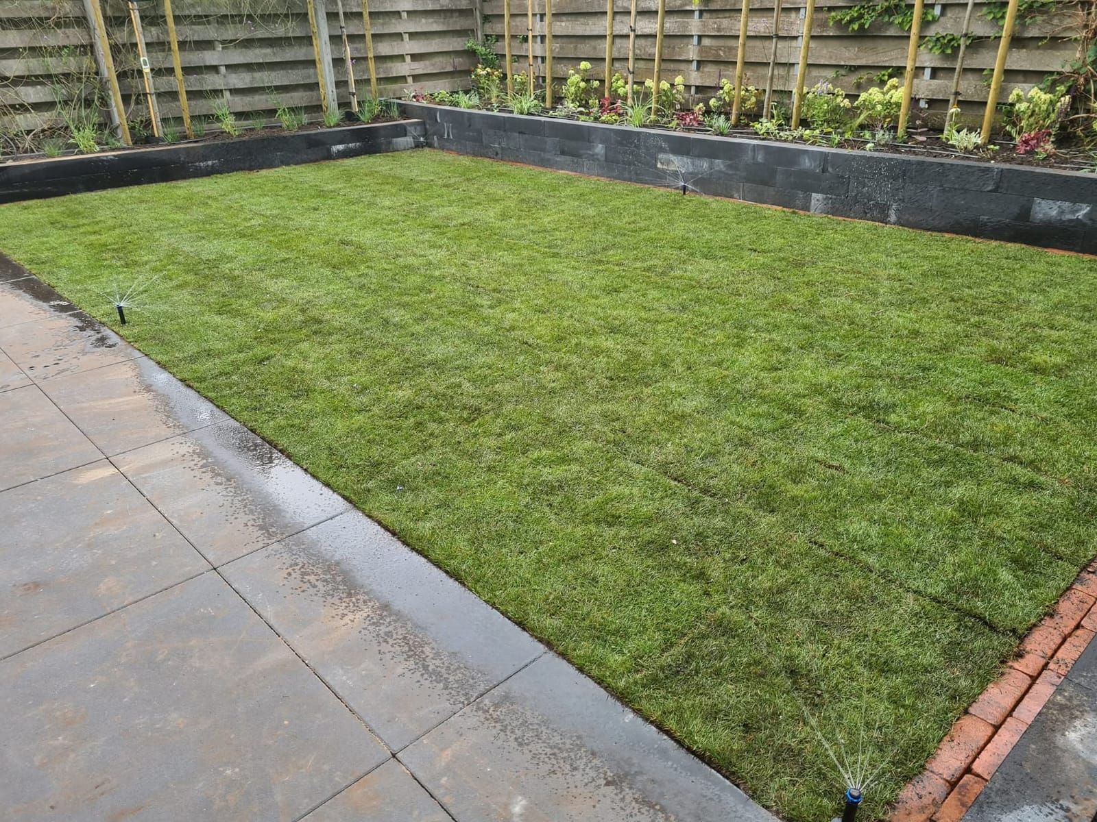 Rechthoekige, met gras begroeide achtertuin, afgebakend door stenen randen, bestrating en een houten schutting.