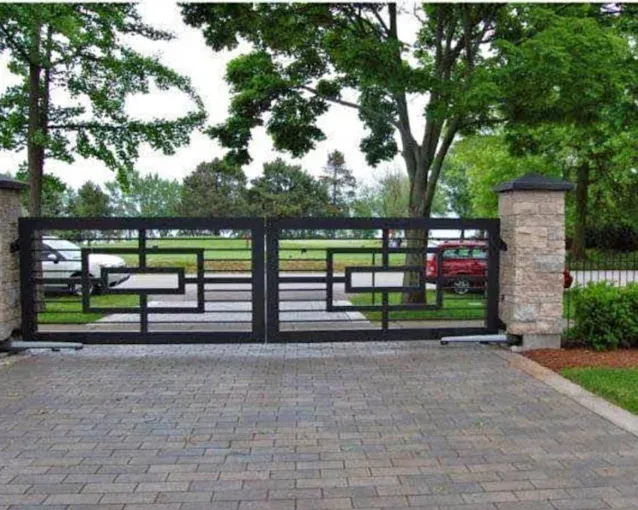 Black metal gate with geometric design on a brick driveway, framed by stone pillars and trees.
