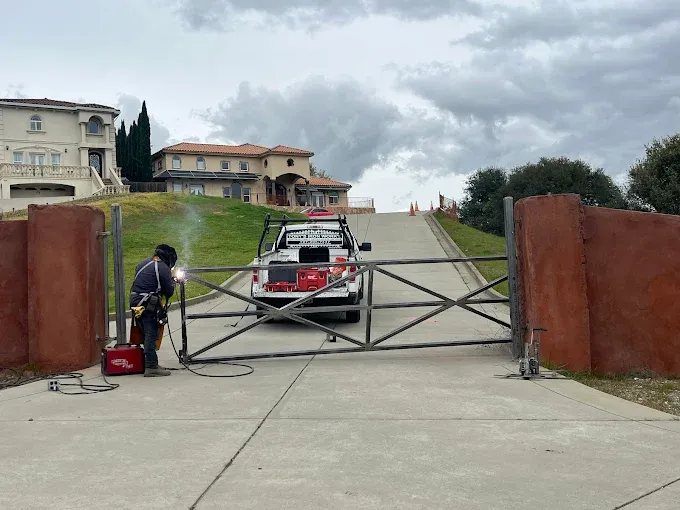 Welder working on a metal gate in front of a residential driveway, truck parked behind the gate, overcast sky.