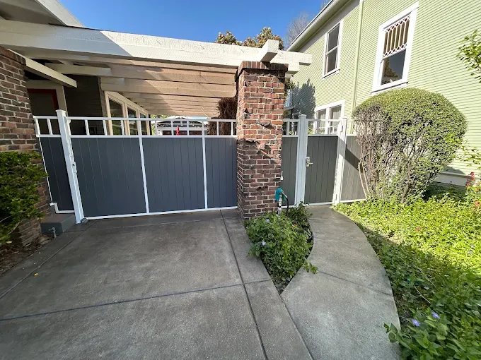 Gray gate with white trim, brick pillar, and a concrete path leading to a green house.