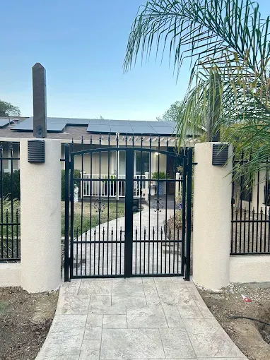 Black wrought-iron gate in beige pillars on a stamped concrete walkway leading to a house with solar panels.
