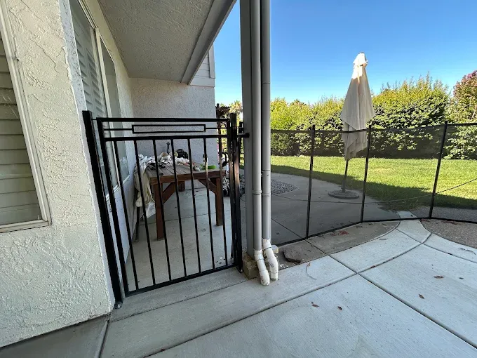 Black metal gate on a patio, leading to a yard with a fence and umbrella on a sunny day.