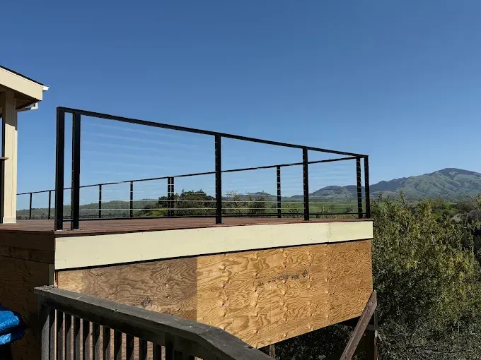 Deck with black railing, clear panels, overlooking a valley under a blue sky.