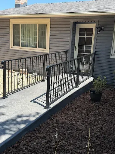 Ramp with black metal railings leading to a door; dark grey siding on the house; sunny outdoor setting.