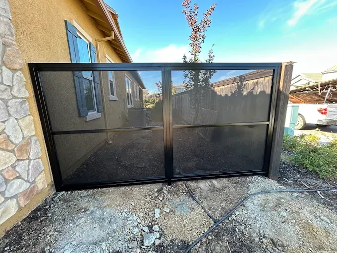 Black metal gate with mesh panels, in front of a tan building and wooden fence, outdoors.