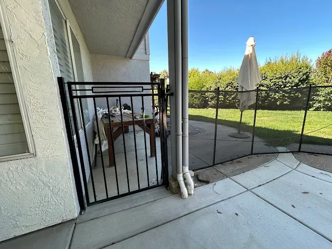 Black gate on patio next to a building, leading to a yard with a fence and umbrella.