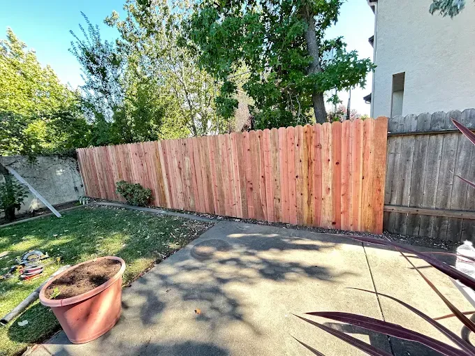 Wooden fence in a backyard, with a concrete patio in the foreground and trees in the background.