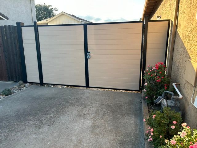 Closed beige driveway gate beside a yellow house wall, with flowers along the right side.