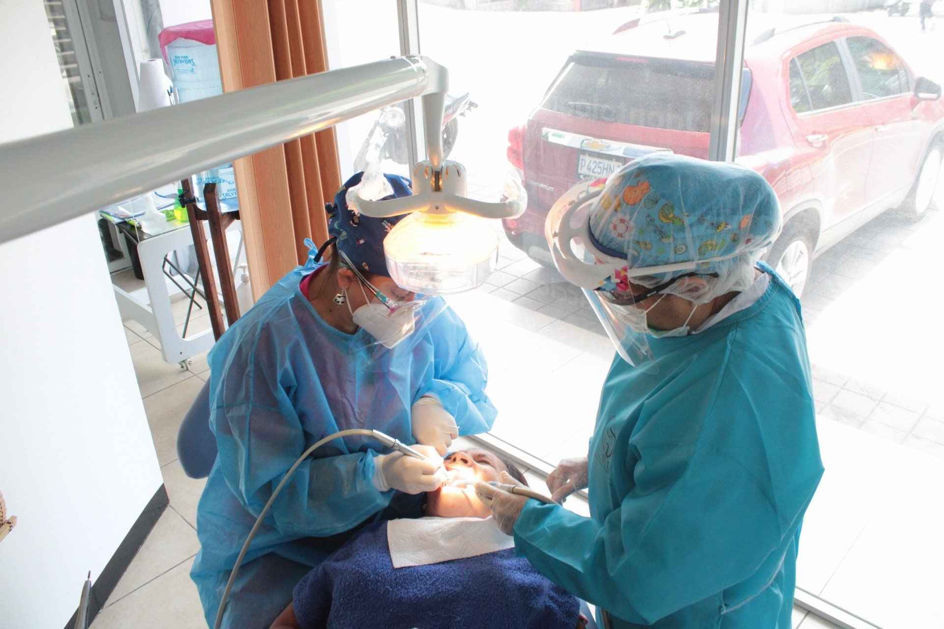 Dentista atendiendo a un paciente en una clínica luminosa, con dos empleados dentales enmascarados y con equipo de protección.