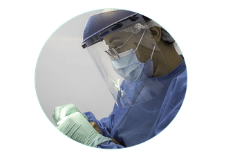 Trabajador sanitario con EPI azul, protector facial y mascarilla, mirando hacia abajo a unas manos enguantadas.