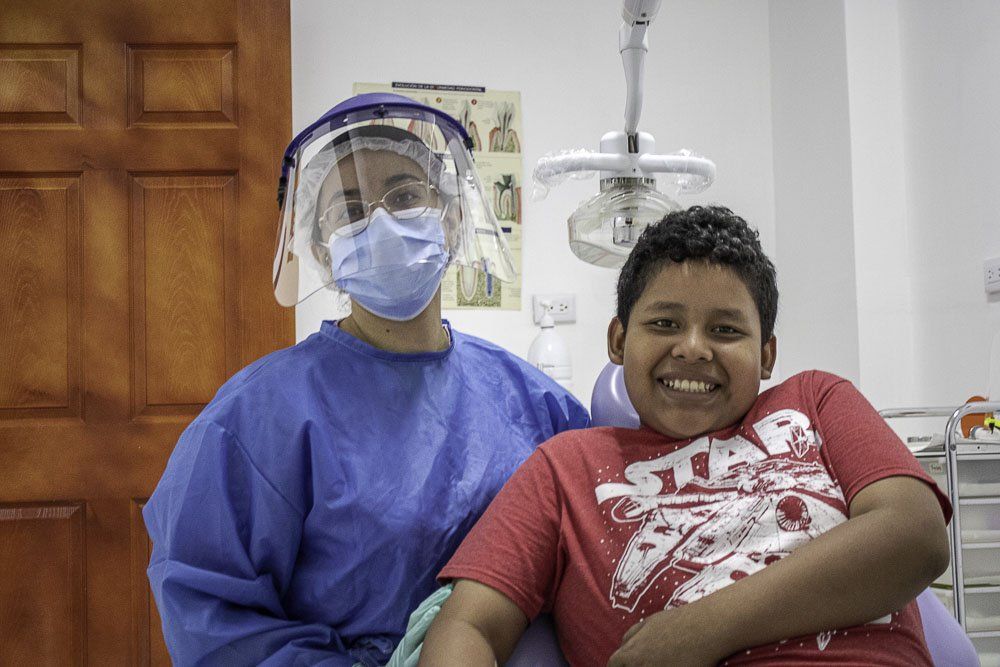 Un trabajador sanitario con traje protector azul y careta facial se sienta junto a un paciente sonriente en una sala de consulta.