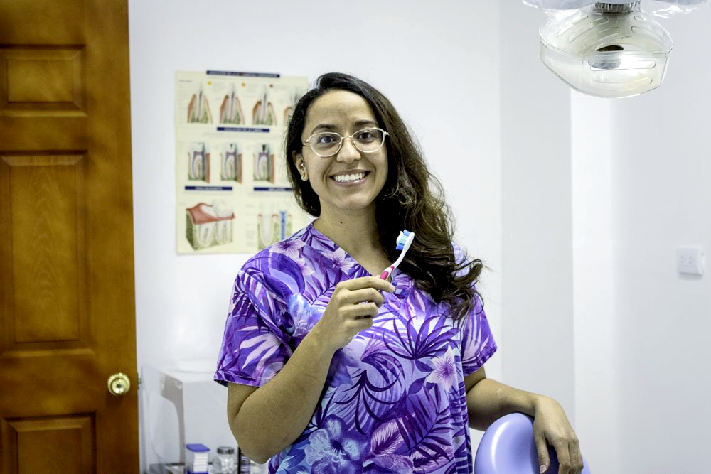 Mujer con blusa morada sonriendo y sosteniendo un cepillo de dientes en un baño o consultorio médico.
