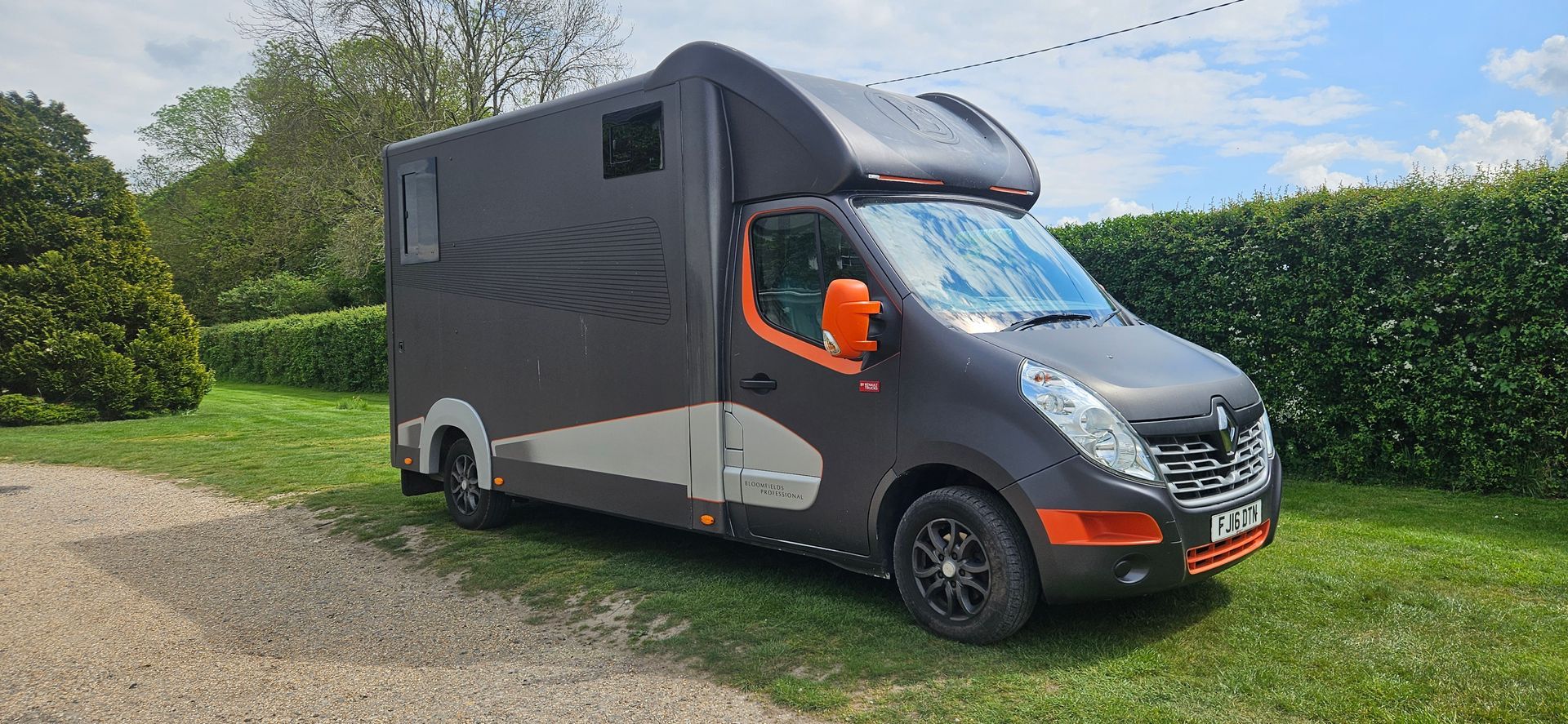 Dark gray delivery van parked on a gravel driveway beside a hedge, with an orange object in the window.