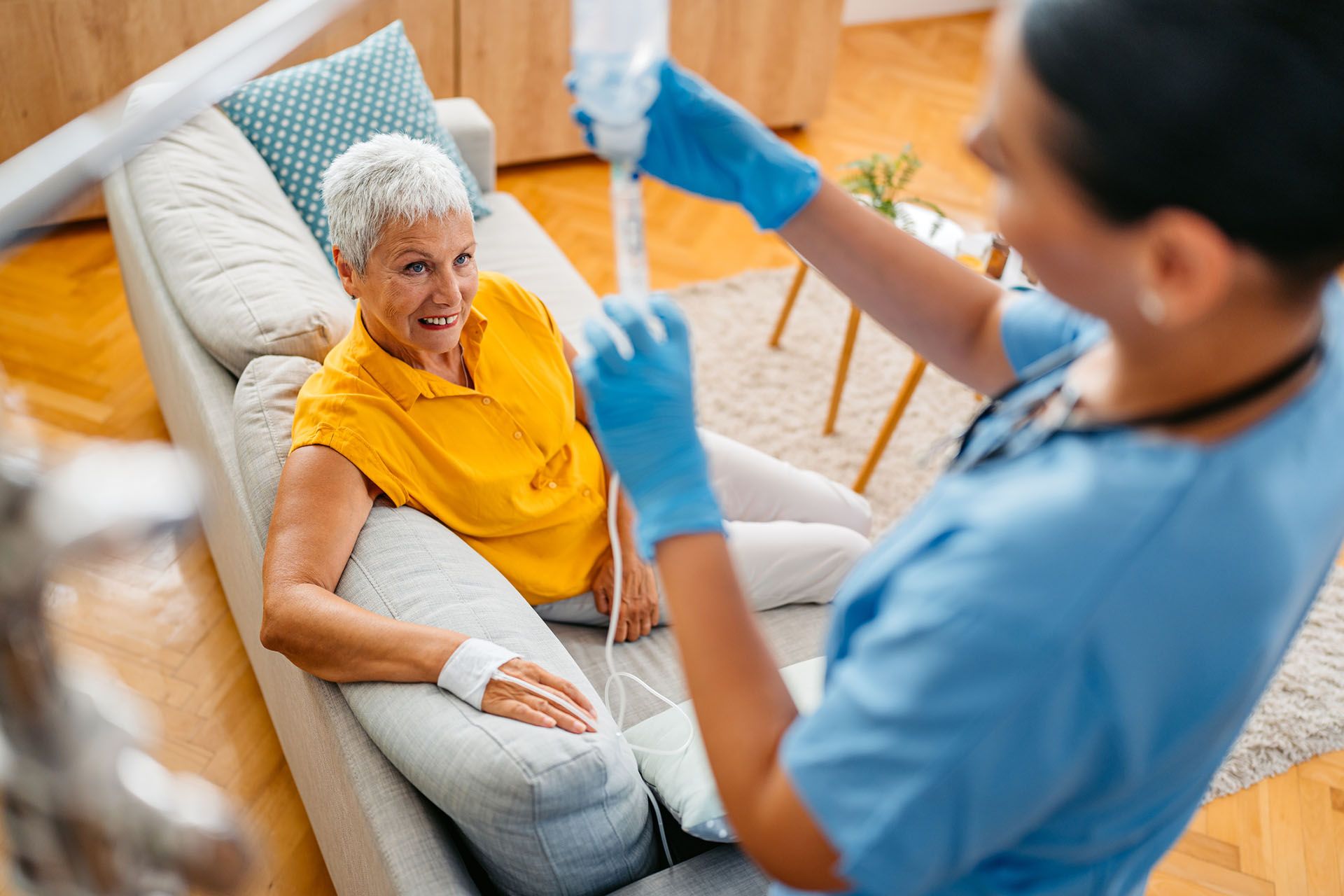 A nurse sets up an IV for an older woman on a couch