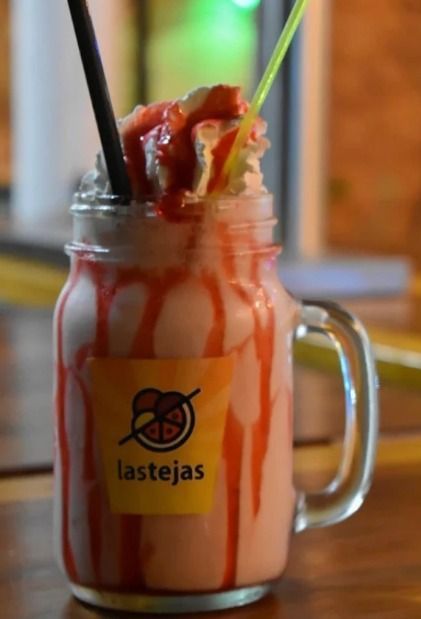 Bebida en tarro de cristal con sirope rojo, nata montada y dos pajitas sobre una mesa de madera; se ve el logo de Lastejas.