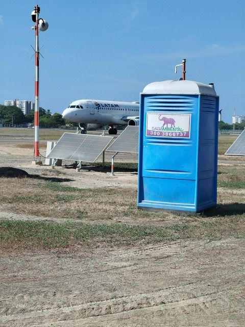 Baño portátil azul en un aeródromo, con un avión estacionado y un marcador rojo y blanco en el fondo.