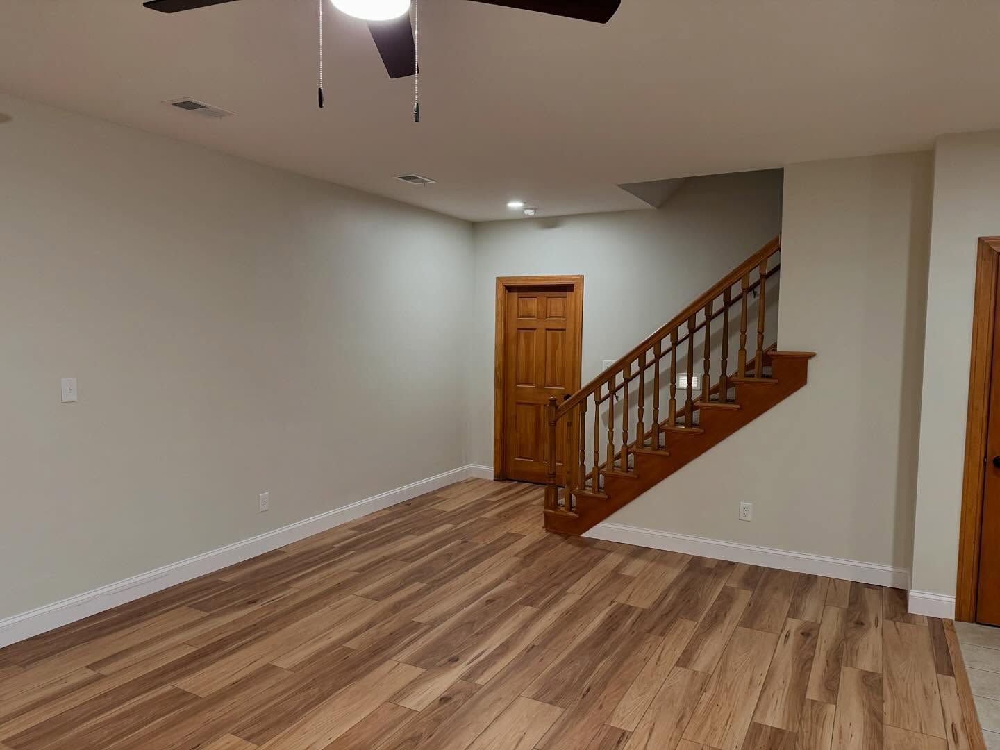 Empty living room with wood floor, beige walls, and a staircase beside a wooden door. Empty living room with wood floor, beige walls, and a staircase beside a wooden door.