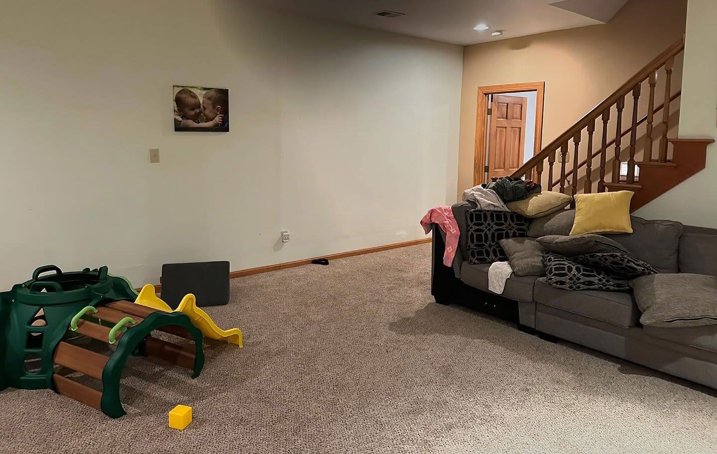 Living room with beige carpet, gray sofa, staircase, and a green plastic toy on the floor. Living room with beige carpet, gray sofa, staircase, and a green plastic toy on the floor.