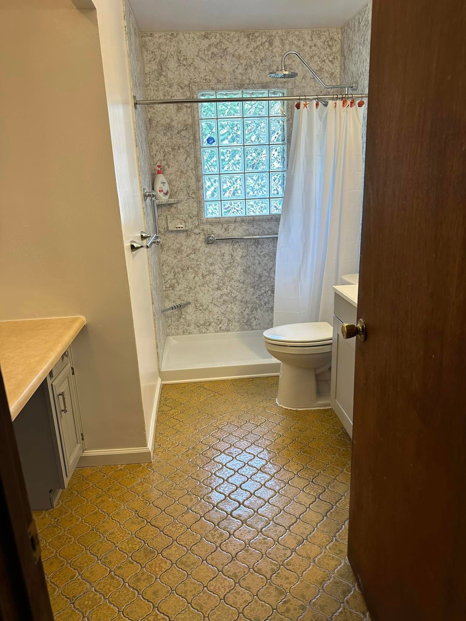Small bathroom with patterned floor, tiled shower area, toilet, and frosted window, viewed from the doorway Small bathroom with patterned floor, tiled shower area, toilet, and frosted window, viewed from the doorway