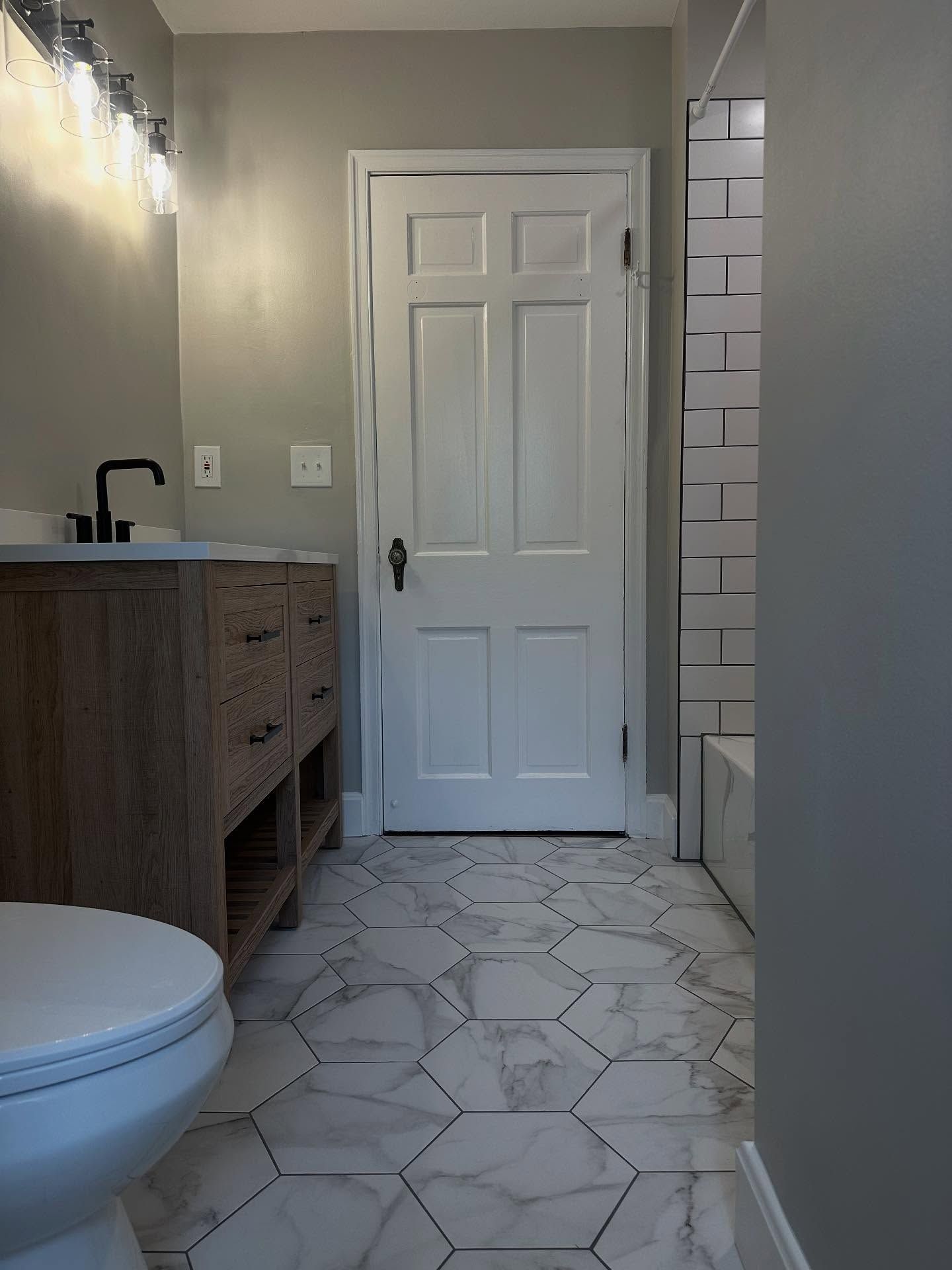 Small bathroom with white door, hex tile floor, gray walls, wood vanity, and toilet in foreground Small bathroom with white door, hex tile floor, gray walls, wood vanity, and toilet in foreground
