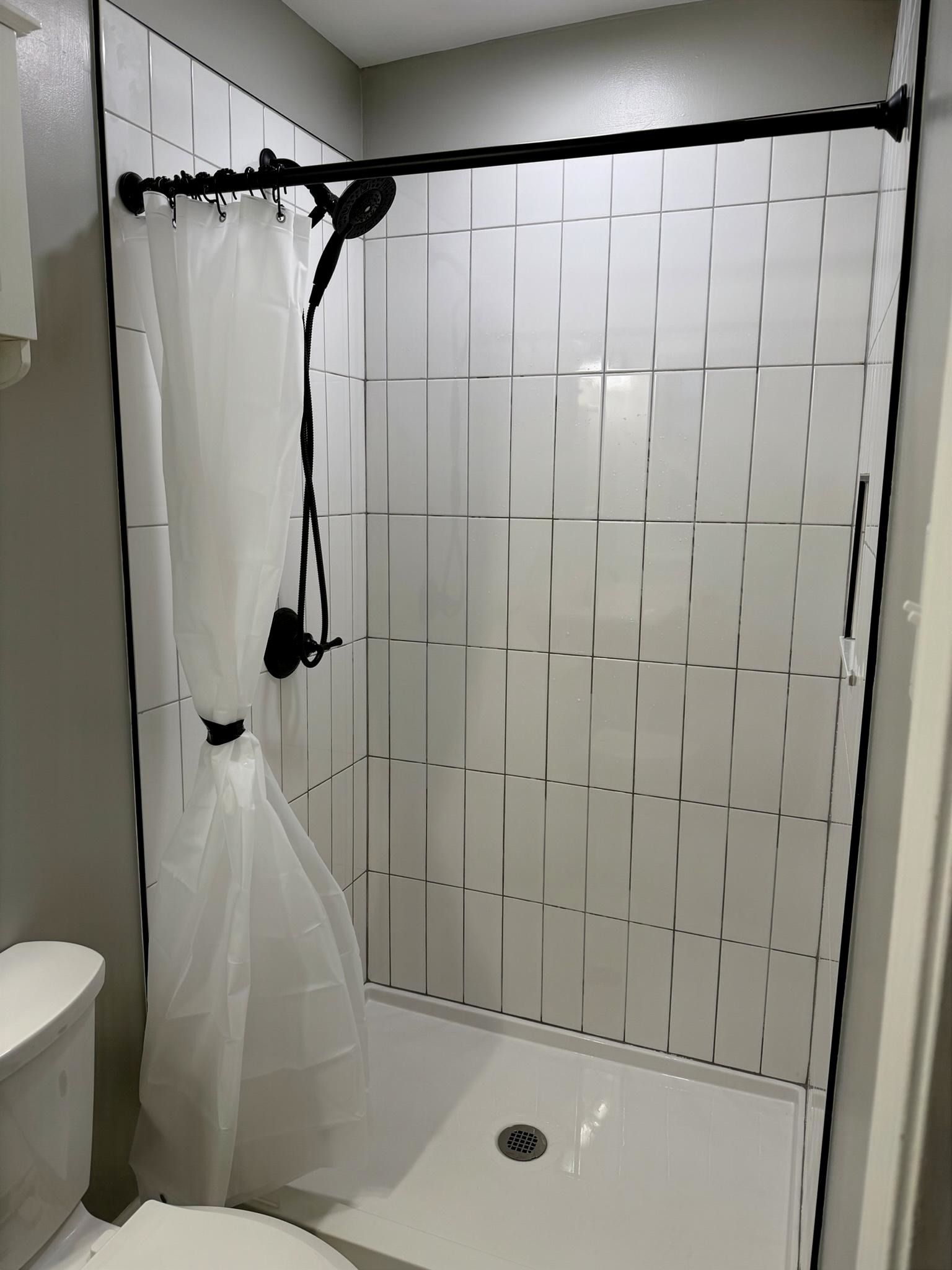 White-tiled shower with a black curtain rod, white curtain, and a showerhead over a small shower base. White-tiled shower with a black curtain rod, white curtain, and a showerhead over a small shower base.