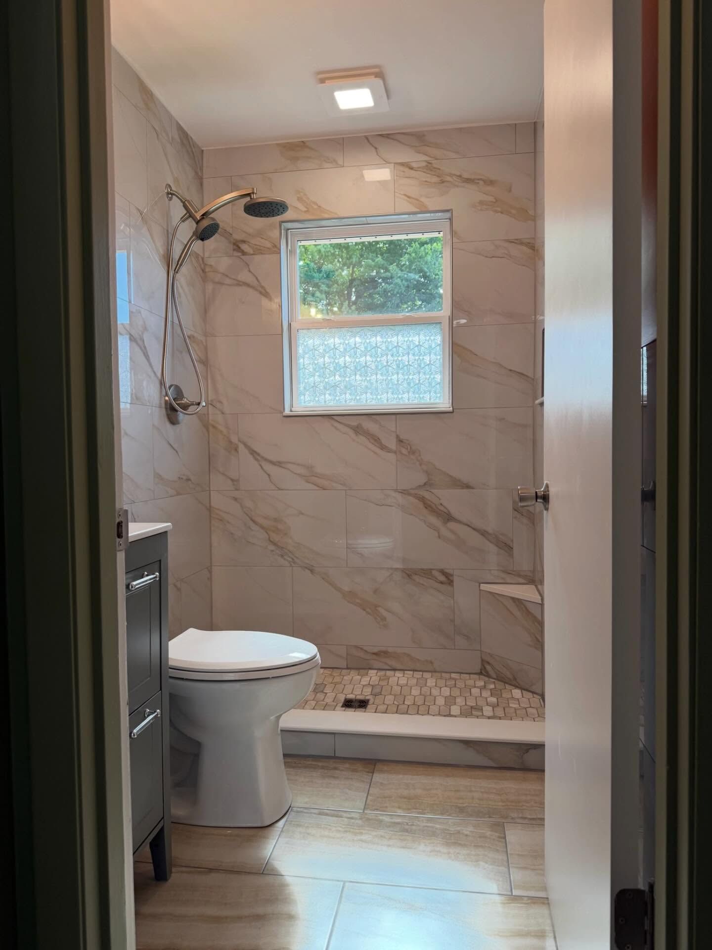 Bathroom with toilet, marble-look tiled walls, shower, and small window with a frosted lower pane. Bathroom with toilet, marble-look tiled walls, shower, and small window with a frosted lower pane.