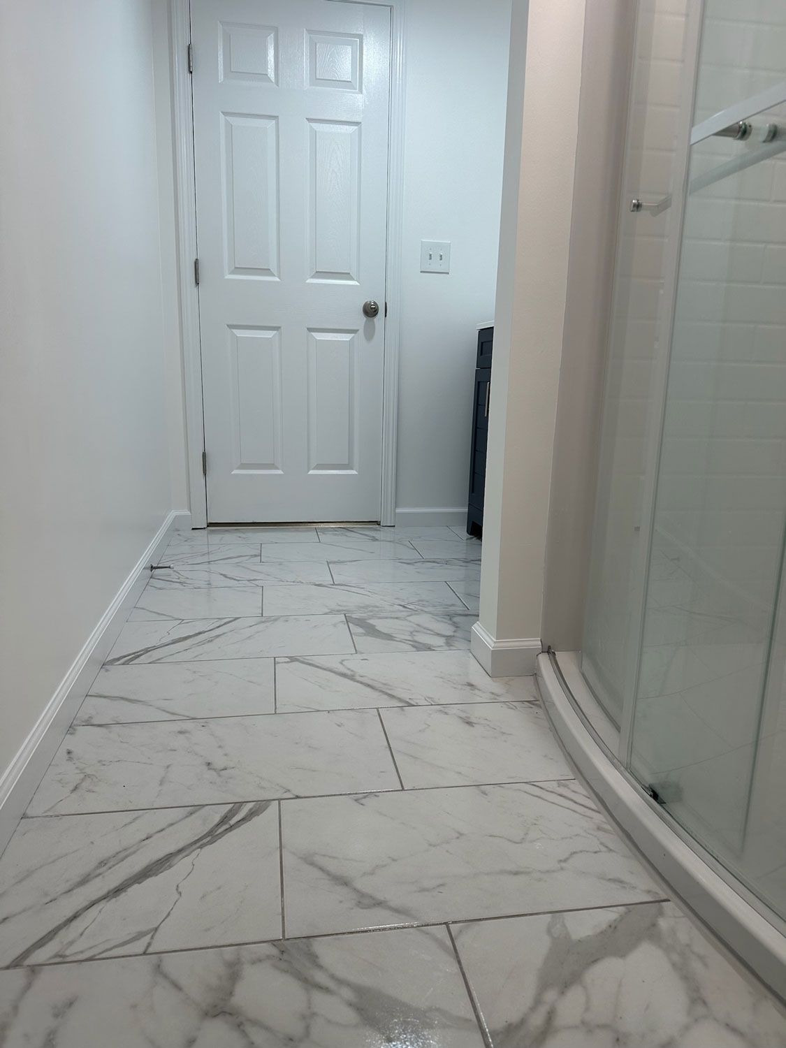 Narrow hallway with white paneled door, marble-pattern tile floor, and glass shower enclosure on the right Narrow hallway with white paneled door, marble-pattern tile floor, and glass shower enclosure on the right